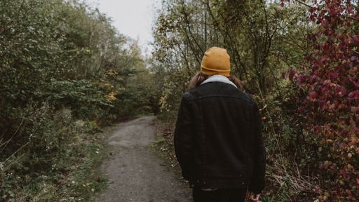A back view of a female wearing a jacket and an orange hat, walking on a narrow dirt road through a forest