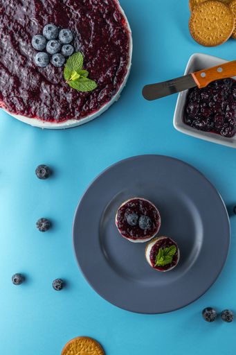 A vertical top view of blueberry cheesecakes with mint leaves, jam, and biscuits on blue surfa