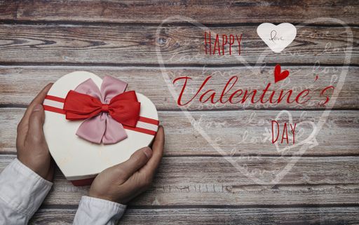 A top view of hands holding a heart-shaped gift box with "Happy Valentine's Day" calligraphy text on wooden table