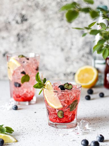 Two fresh summer iced drinks garnished with lemon, mint, and blueberries, a vertical shot