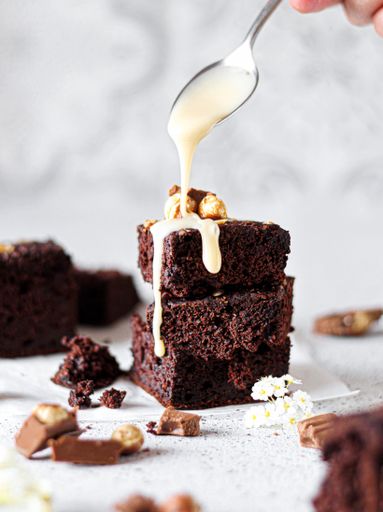 A closeup shot of a person pouring white chocolate cream over chocolate brownies, served with chocolate and caramel on a bright background