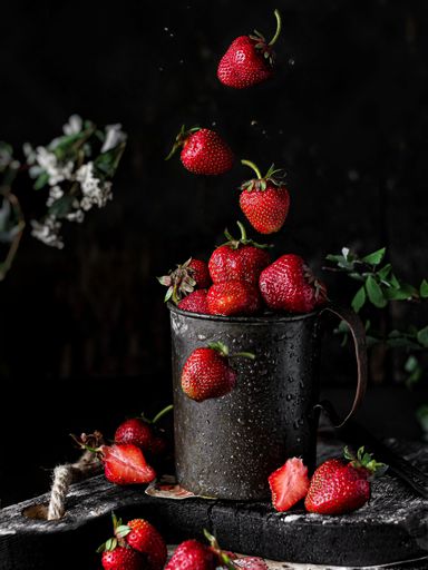 A closeup shot of a wet vintage cup with strawberries, on a wooden board, surrounded by falling strawberries, on a black background