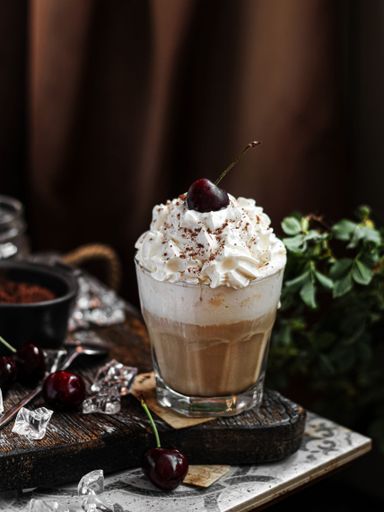 A vertical shot of ice coffee with white cream in glass cup on table with cherry next to it