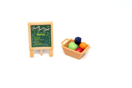 A basket of mini plastic toy fruit next to a juice menu board with a concept of heathy eating. doll house miniature fruit. a close up photo