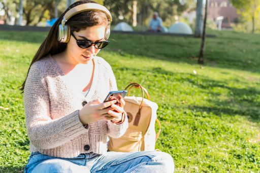 A smiling woman with headphones listening to music and using a smartphone sitting in a park