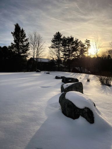 A vertical shot of rocks covered in snow in a field with a house and trees on a sunny day