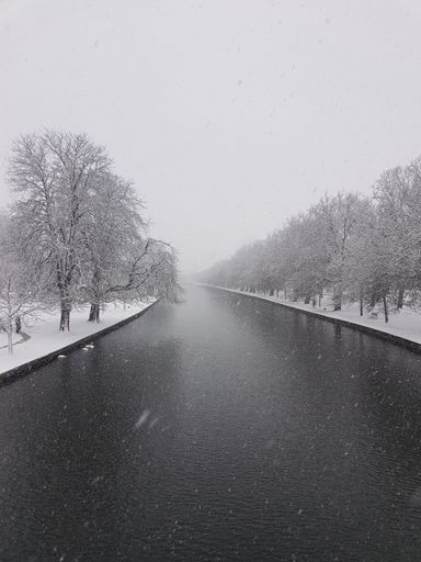 A vertical shot of an urban river surrounded by banks covered with snow in winter