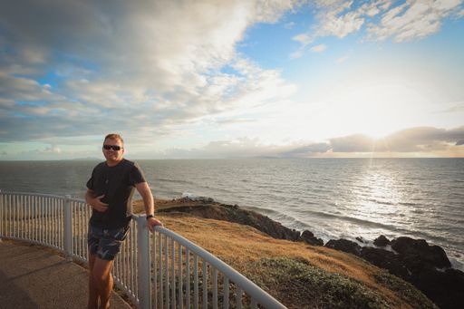 A man standing near metallic fences surrounded by greenery and the sea during the sunset