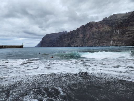 A scenic view of people swimming in ocean waves against rocky cliffs on a cloudy day