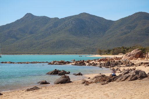 A beach covered in rocks surrounded by the sea and hills covered in forests under sunlight