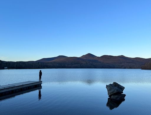 A view of a person standing on a wooden pier and a rock in calm water reflecting the clear blue sky with a mountain in the background