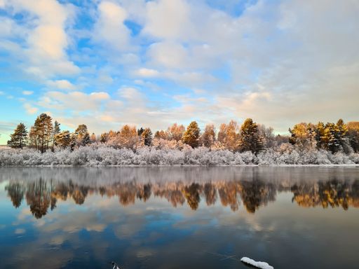 A breathtaking view of a calm lake's surface and a reflection of the lake's coast, at sunset on a winter day, with clouds in the blue sky