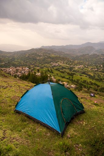 A vertical of a blue and green tent on top of a green hill on a cloudy day