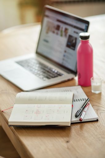 A selective focus shot of a laptop, thermos flask, calendar and pens on a textbook
