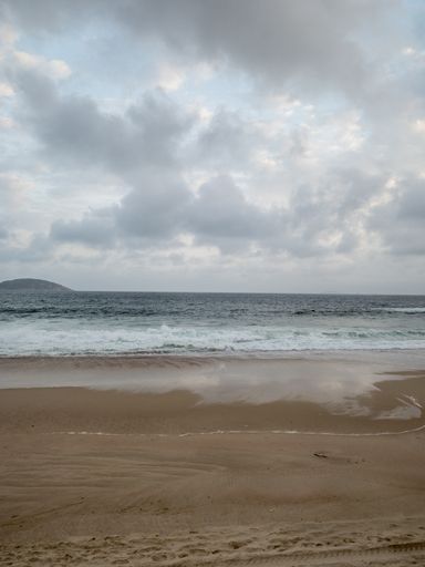 A vertical shot of a sandy beach and wavy sea under cloudy sky