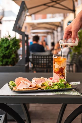A vertical shot of a plate with ham bagels and greens near iced coffee with a hand pouring milk