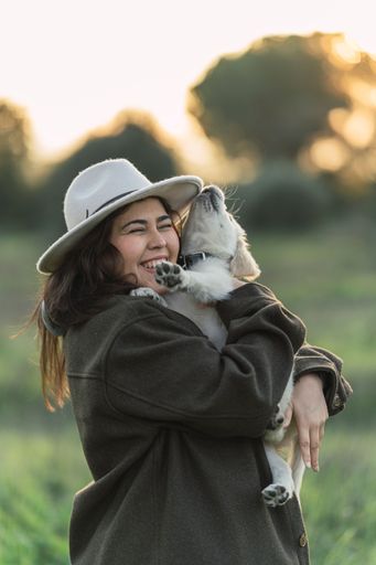 A young Caucasian woman wearing a hat standing in a grassy field, lovingly holding her small Golden Retriever dog at sunset
