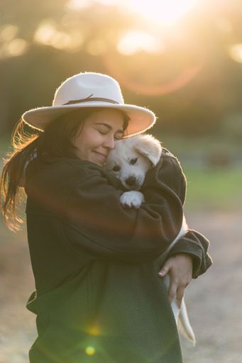 A young Caucasian woman wearing a hat standing in a grassy field, lovingly holding her small Golden Retriever dog at sunset