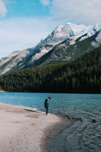 A vertical shot of a boy standing on the shore beside the body of water near the mountains