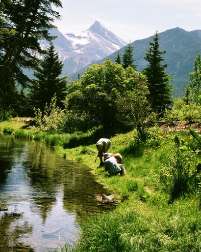 A vertical shot of two people on the grassy shore of the creek near the mountains