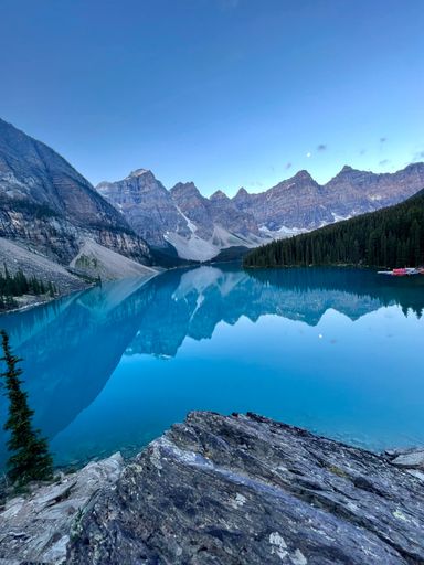 A vertical shot of a clear blue lake surrounded by a forest and a mountain range in the background