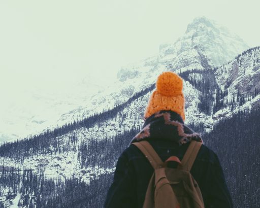 A vertical shot of a person in a yellow hat and with a backpack standing in front of the snowy mountains