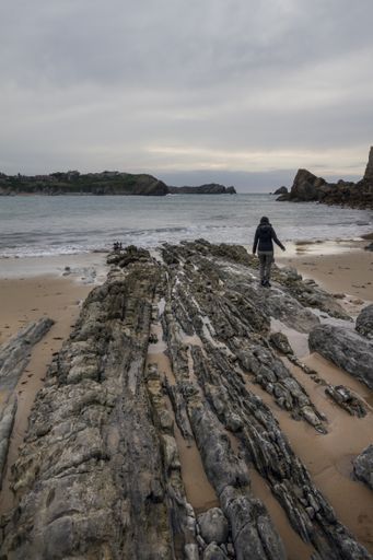 A vertical shot of a person walking on a cliffy seashore near a wavy sea under a cloudy sky