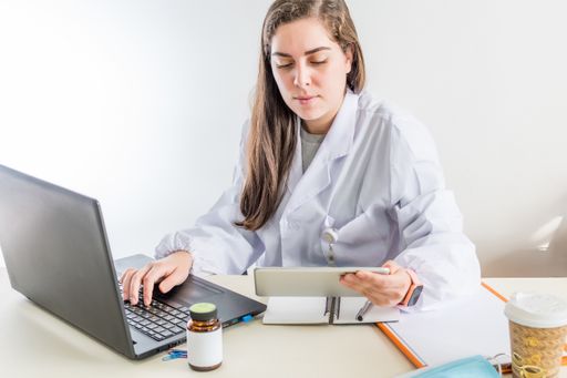 A young female doctor sitting in her consulting room consulting data on a tablet while writing a report, medical diagnosis