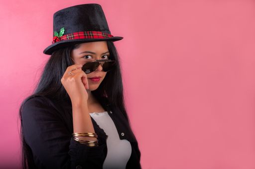 A beautiful Latin female in sunglasses and a hat standing next to a pink wall