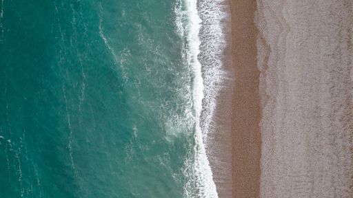The image shows a drone shot of the beach of Etretat in Normandy. The blue water in combination with the sand and stone beach is a beautiful contrast.