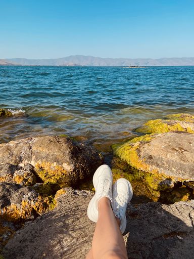 A vertical shot of female legs on the coastline rocks with the calm blue sea on the background