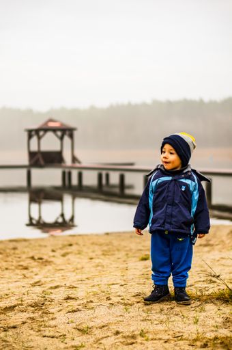 Young small boy wearing warm clothes standing on sand close by a lake on a cold day with mist in the winter season.