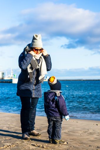 Woman and child wearing warm winter clothes standing on sand close by the sea on a cold day.