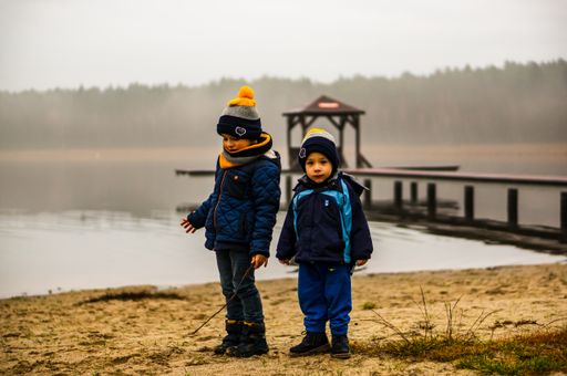 Two small boys standing on sand close by the water by a lake with wooden pier on a cold and misty day.