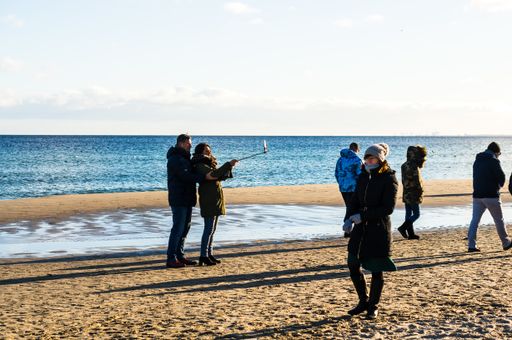 Group of people walking on sand at a beach on a cold day. Couple taking a selfie close by the water.