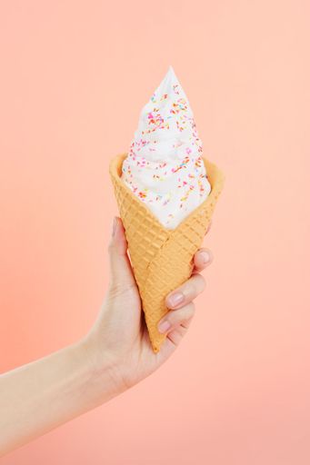 A female's hand holding delicious soft ice cream with sprinkles in a crispy waffle cone on a pink background
