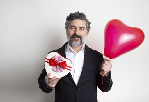 A bearded male in suit holding a red heart-shaped box with a balloon on a white background