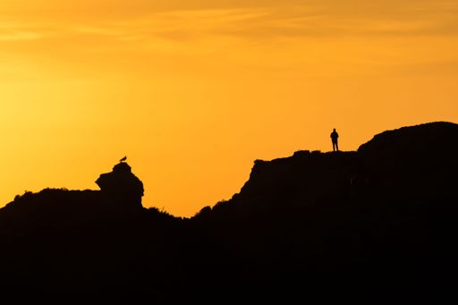 A person on massive rocks silhouetted against the backdrop of an orange sky at sunset.