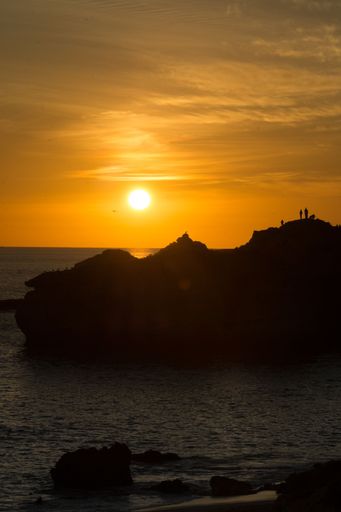 The people on massive rocks silhouetted against the backdrop of an orange sky at sunset.