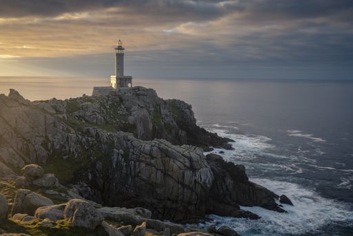 The Punta Nariga Lighthouse on the cliff under the sunrays piercing through the cloudy sky in Spain