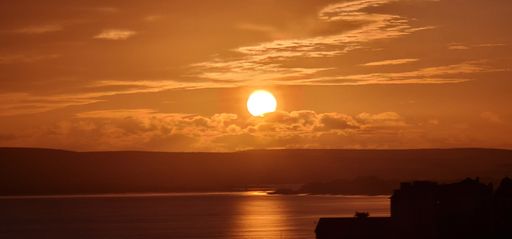 A panoramic view of the sunset scene over mountains around the sea with a silhouette view of the coastal buildings in the foreground