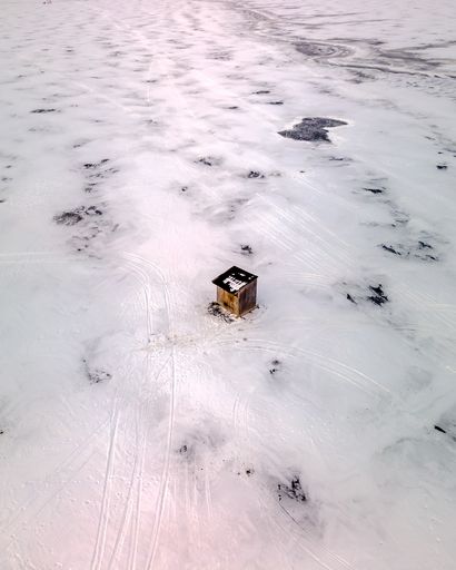 An aerial view of a wooden small hut in the middle of a frozen snow-covered field with ski tracks