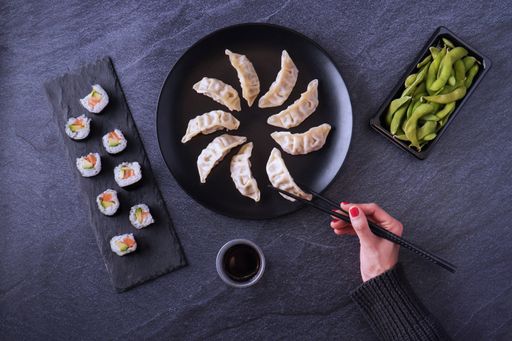 A top view shot of a female hand taking chicken gyoza, the salmon and avocado makis, edamame beans, and soy sauce served on the table