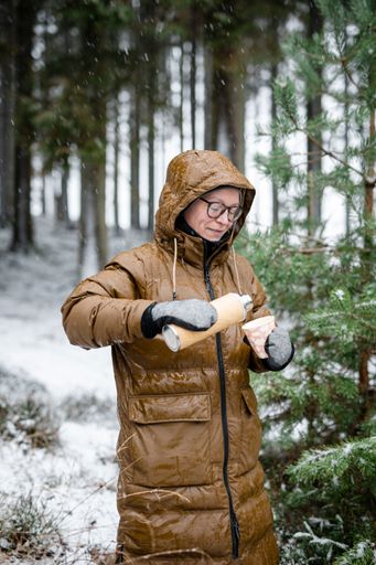 A vertical shot of a happy female in a winter coat pouring a hot drink from an insulated bottle under snowfall in a forest