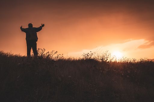 A silhouette closeup of a male standing with open arms in the meadow during the sunset