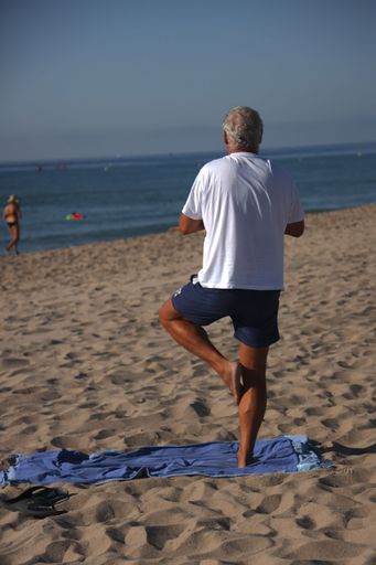 A vertical shot of a man standing on a one leg on a sandy beach on a sunny day