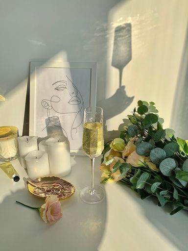 A vertical shot of a glass of champagne, bouquet of flowers, candles, and a picture on a white table