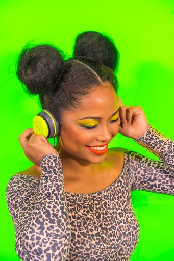 A closeup portrait of a pretty Hispanic woman with bright makeup and two side bun hairstyle listening to music with headphones