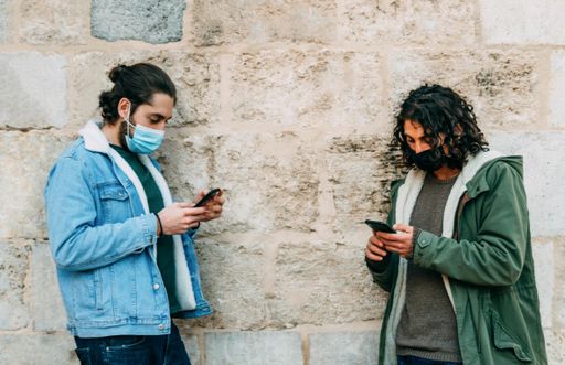 Two young Caucasian males with medical masks leaning on a wall and texting on the phone