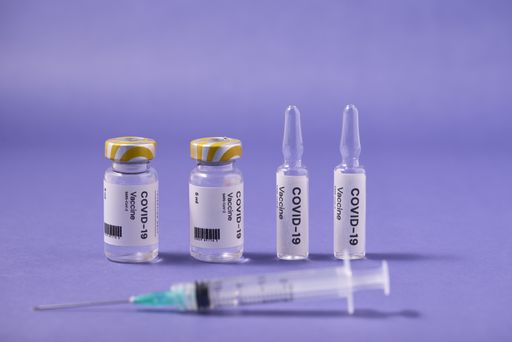 A closeup of a syringe needle with the COVID-19 vaccine ampoules on a purple surface